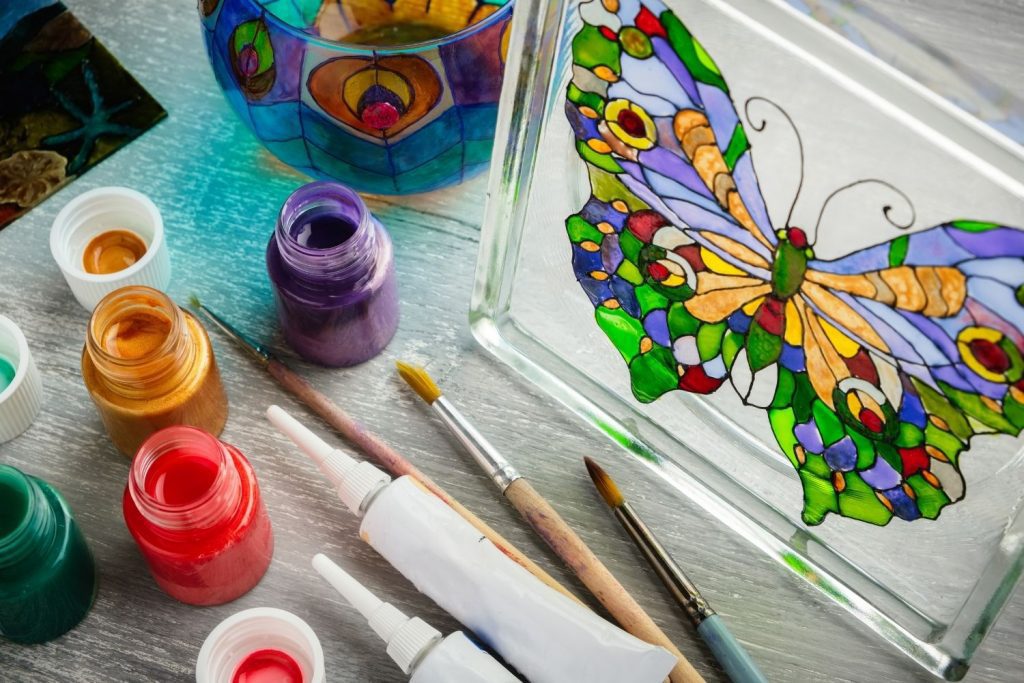 Butterfly painted on glass with paint bottles and brushes on table. Stained glass art for kids.